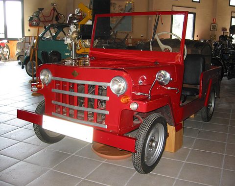 Red jeep-style open microcar with a bar grille and a raised windscreen, parked on a tiled museum floor. Opens a larger view