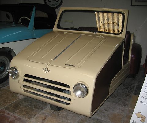 Cream three-wheeler with dark side panels, a slatted grille and two round headlamps, in a museum.