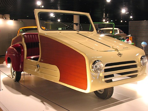 A small open cream three-wheeler with wood-grain side panels, red leather seats and a slatted grille under spotlights. Opens a larger view