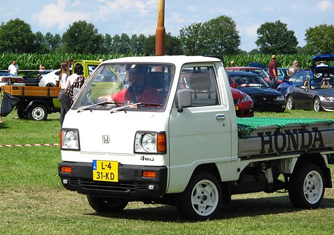 White cab-over pickup with a green cover lettered HONDA, on grass with visitors behind. Opens a larger view