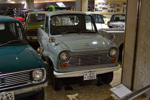 Pale blue-grey small pickup with a 1965 plate, a teal car in the foreground at left. Opens a larger view