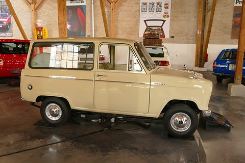 Side view of a beige small van in a brick hall with wooden beams, a red car behind. Opens a larger view