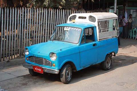 Bright blue small pickup with a white cover, parked by a slatted wooden fence. Opens a larger view
