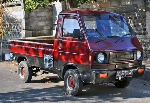 Red cab-over pickup with a black bumper, parked by a stone wall in dappled sun. Opens a larger view