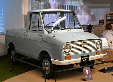 Pale blue-grey small pickup with a slatted grille on a museum floor, a large photo mural behind. Opens a larger view