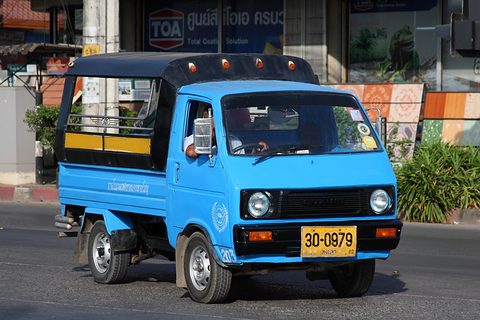 Blue cab-over pickup with a roofed bed and a yellow plate, in a street. Opens a larger view