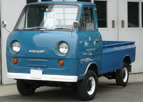 Blue cab-over pickup with round headlamps parked in front of a white building. Opens a larger view