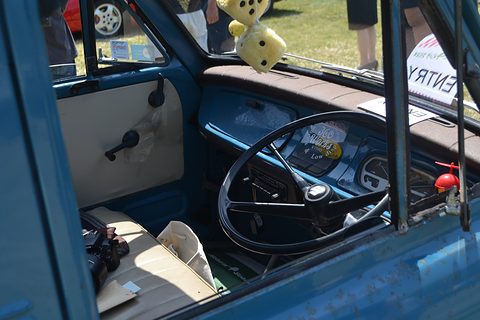 Through an open door: a large black wheel, a blue dashboard and a cream seat, fluffy dice hanging. Opens a larger view