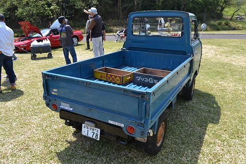 Rear three-quarter of a blue cab-over pickup with wooden crates in the bed, on grass. Opens a larger view