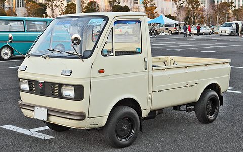 Cream cab-over pickup with square headlamps on a car park, marquees behind. Opens a larger view