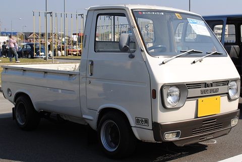 Pale grey cab-over pickup with a yellow plate, flagpoles and cars behind. Opens a larger view