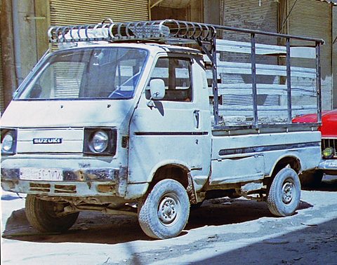 White battered cab-over pickup with a tall metal rack over its bed, parked in strong sun. Opens a larger view