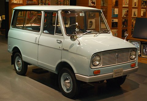 Pale blue small van with a short bonnet on a dark floor, bookshelves behind. Opens a larger view