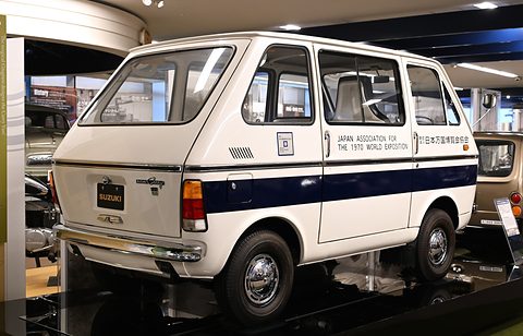 Rear three-quarter of a white van with a navy stripe and SUZUKI plate on a dark plinth. Opens a larger view