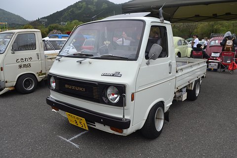 White cab-over pickup with square headlamps and a yellow plate in a busy car park. Opens a larger view
