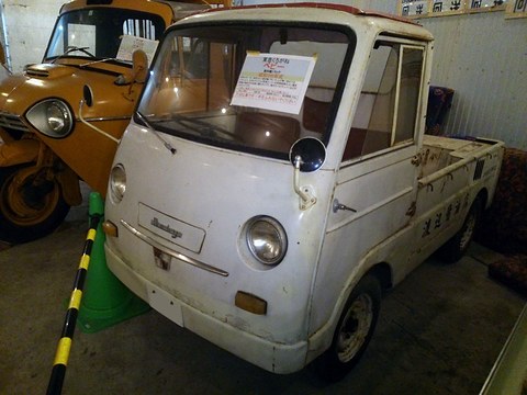 White small cab-over pickup with a sign in its windscreen, an orange three-wheeler beside it. Opens a larger view
