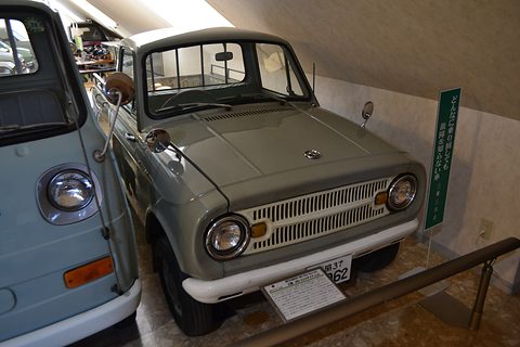 Grey-green small pickup with a slotted grille behind a rail, a blue van at left. Opens a larger view
