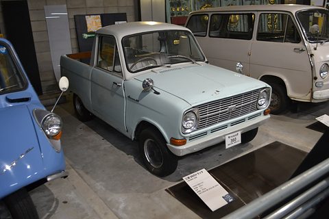 Pale green small pickup on a dark floor with a placard, a white van behind. Opens a larger view