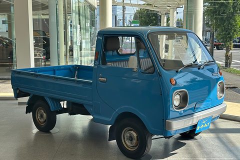 Blue cab-over pickup with round headlamps on a paved forecourt. Opens a larger view