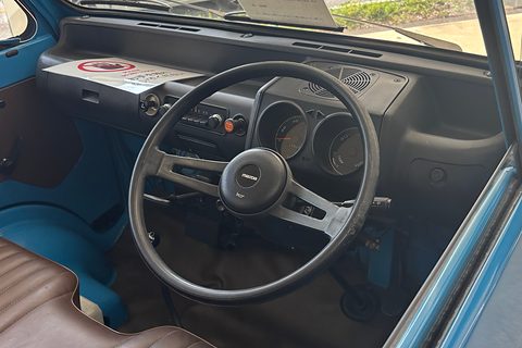 Inside a small truck cab: a black three-spoke wheel, round dials and a brown bench seat. Opens a larger view