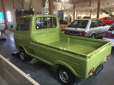 Rear three-quarter of an olive-green cab-over pickup on a dark floor, other cars behind. Opens a larger view