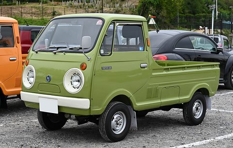 Bright olive cab-over pickup with round headlamps on gravel, an orange truck beside it. Opens a larger view