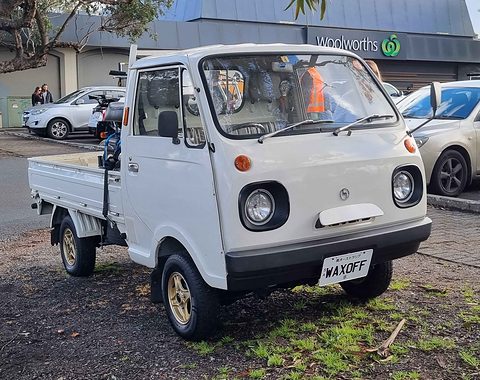 White cab-over pickup with a custom plate on a gravel verge in front of shops. Opens a larger view