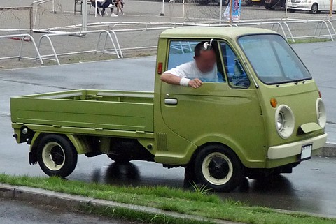 Olive-green cab-over pickup, its driver leaning out of the window, barriers behind. Opens a larger view