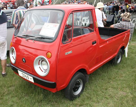 Red cab-over pickup on grass with a 1969 plate, a crowd behind. Opens a larger view