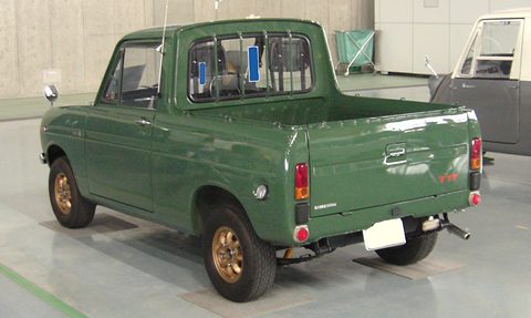 Rear three-quarter of a dark green small pickup with gold wheels, in a hall. Opens a larger view