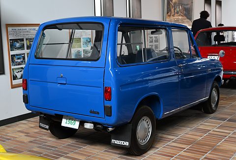 Rear three-quarter of a bright blue small van on a tiled floor with framed pictures behind. Opens a larger view
