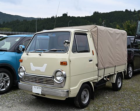 Cream cab-over pickup with a beige canvas cover over its bed, hills behind. Opens a larger view