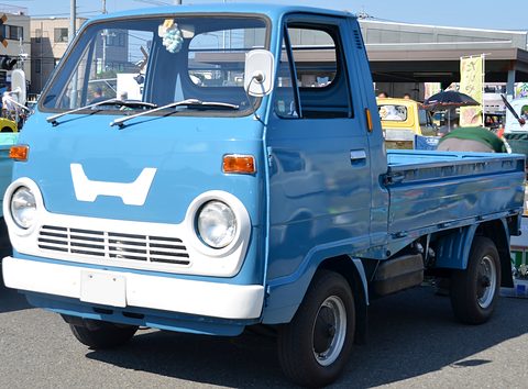 Blue cab-over pickup with a white H panel on its nose, on a car park. Opens a larger view