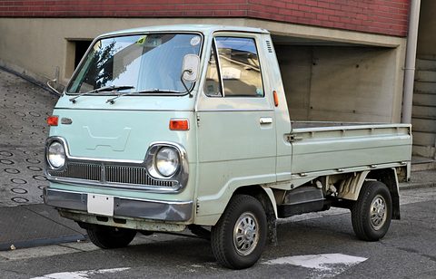 Pale green cab-over pickup with a slotted grille, parked by a garage. Opens a larger view