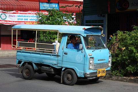 Light blue cab-over pickup with a roofed passenger bed in a street, its driver in the cab. Opens a larger view