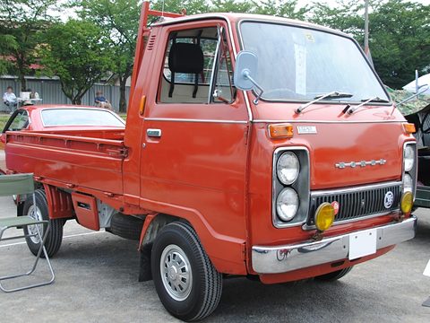 Red cab-over pickup with twin headlamps and a yellow fog lamp, trees behind. Opens a larger view
