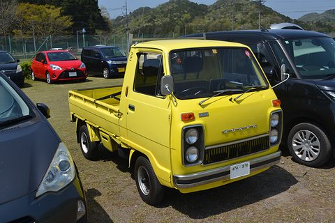 Yellow cab-over pickup between dark modern cars on a gravel car park. Opens a larger view