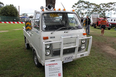 White cab-over pickup with a chrome bull bar and amber roof lamps, on grass. Opens a larger view