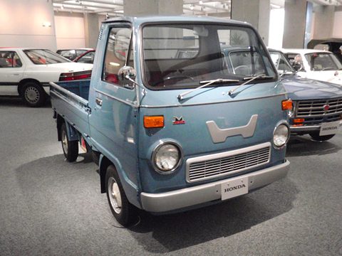 Blue-grey cab-over pickup with a large white H panel on its nose, on a grey floor.