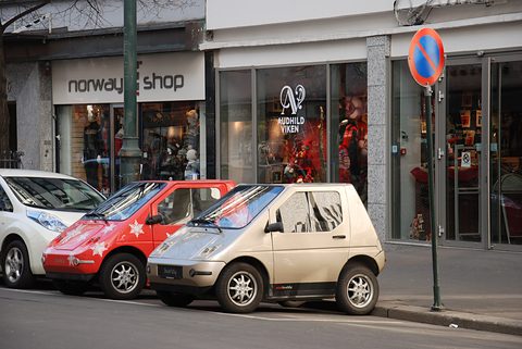 Three small electric cars, white, red with snowflakes and cream, parked nose to tail outside shop windows. Opens a larger view