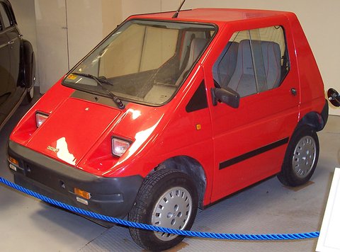 Red angular two-seat electric microcar with grey bumpers behind a blue rope on a museum floor.