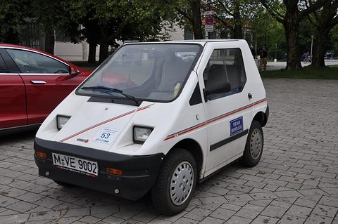 White angular microcar with a red pinstripe and number 53 on its nose, parked on cobbles beside a red car. Opens a larger view