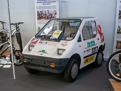 White angular microcar covered in sponsor stickers on a show floor between bicycles and display panels. Opens a larger view