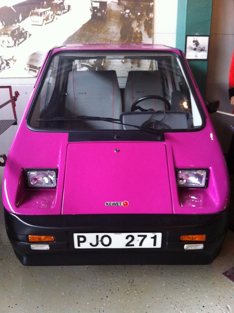 Bright pink angular microcar with black bumpers and a Swedish plate on a museum floor, photographs behind. Opens a larger view