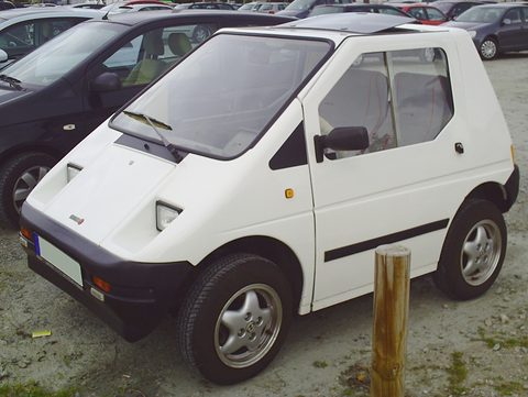 White angular two-seat microcar with black bumpers and alloy wheels on gravel beside a wooden post. Opens a larger view