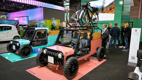 Pink and black open boxy microcar with chunky tyres and a bicycle on its roof rack, a blue one behind. Opens a larger view