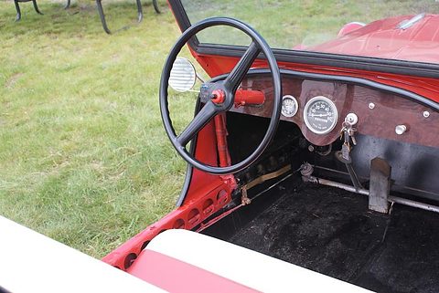 Big black steering wheel over a red dash with a single round speedometer, a small gauge, switches and keys, a pedal on the black floor. Opens a larger view
