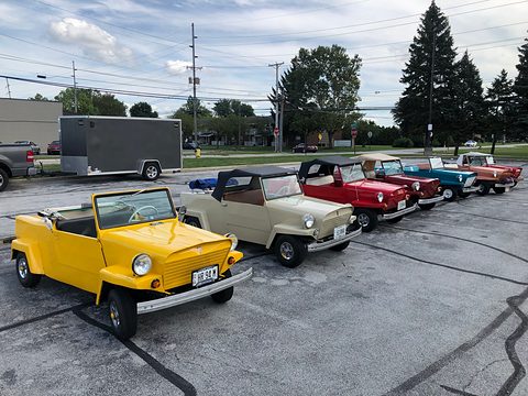 Six small open two-seaters in yellow, beige, red and blue lined up across a car park under a cloudy sky, trees behind.