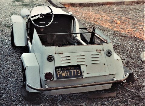 High rear view of an off-white open two-seater with louvres on its engine cover, small round tail lamps and a California plate, the steering wheel beyond. Opens a larger view