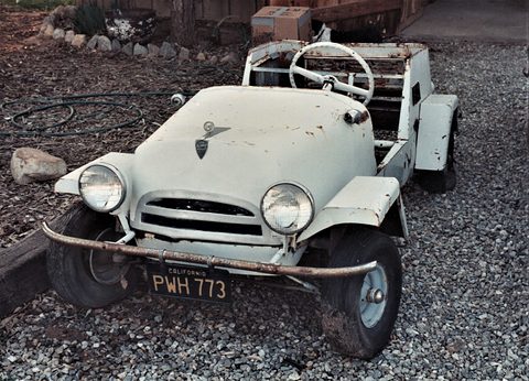 Off-white open two-seater with round headlamps, a chrome grille surround and a thin front bumper, parked on gravel with a large steering wheel visible.